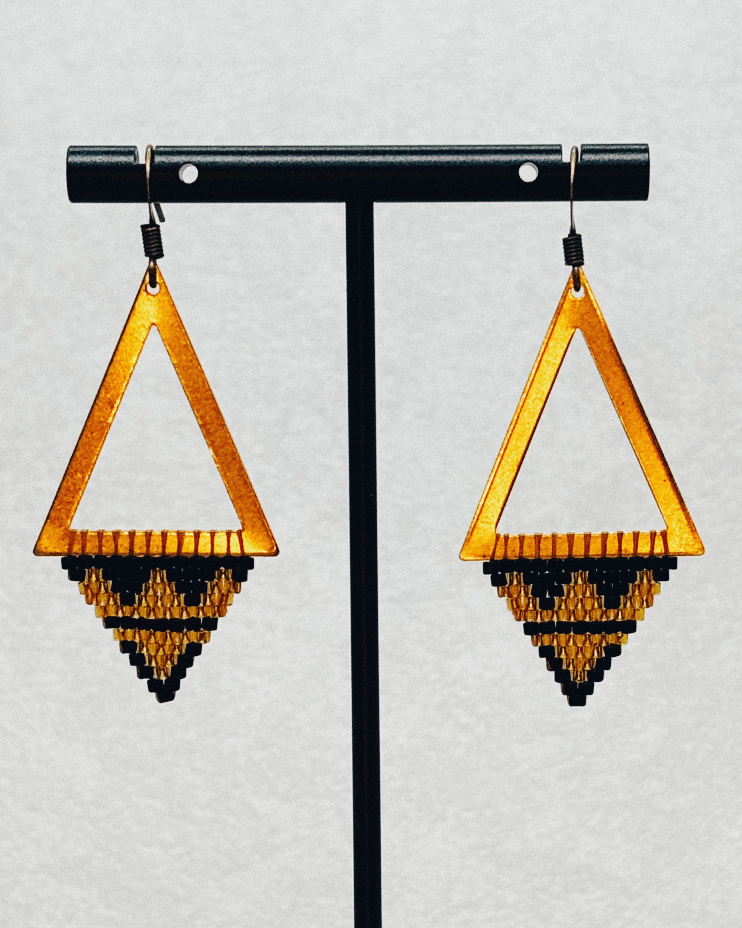 Bronze Triangle Dangles with open bronze frames and handcrafted black and gold geometric bead patterns hanging on display stand